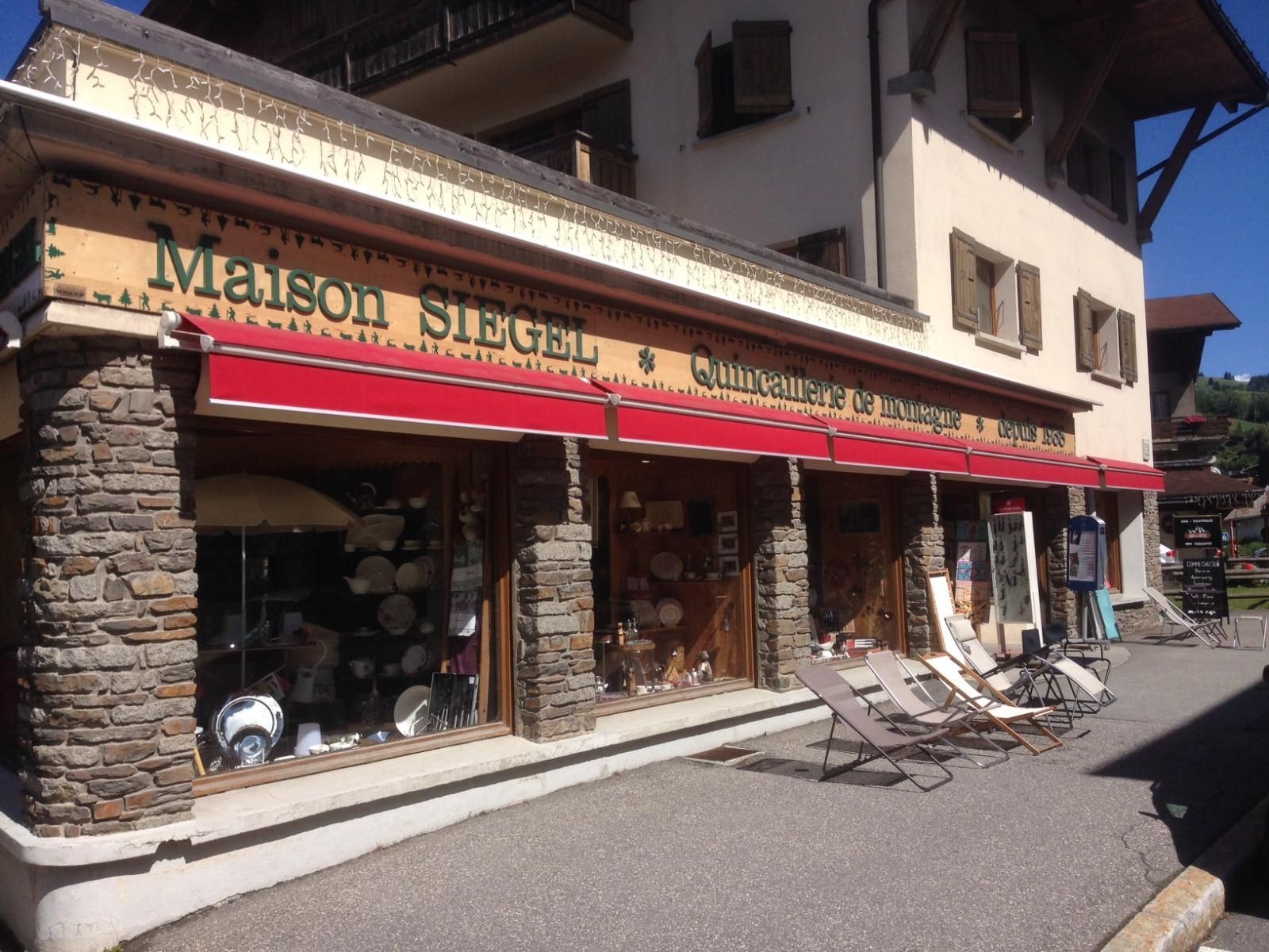 Vue de rue d'une façade rustique avec un auvent rouge et une vitrine extérieure dans une rue ensoleillée d'un village alpin.