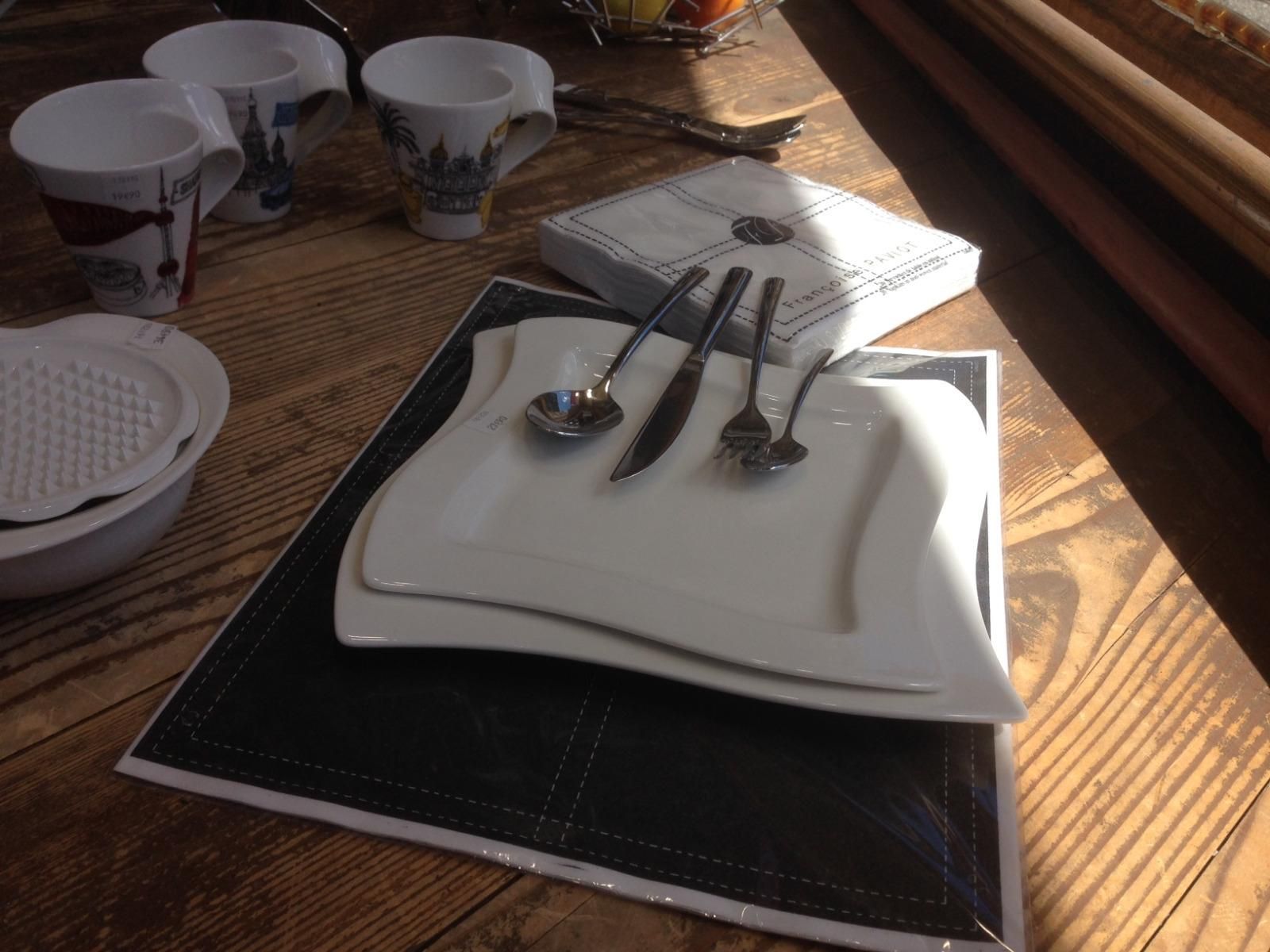 Dresser la table à manger avec des assiettes blanches empilées, des couverts et des tasses sur une nappe tissée près d'une fenêtre ensoleillée.