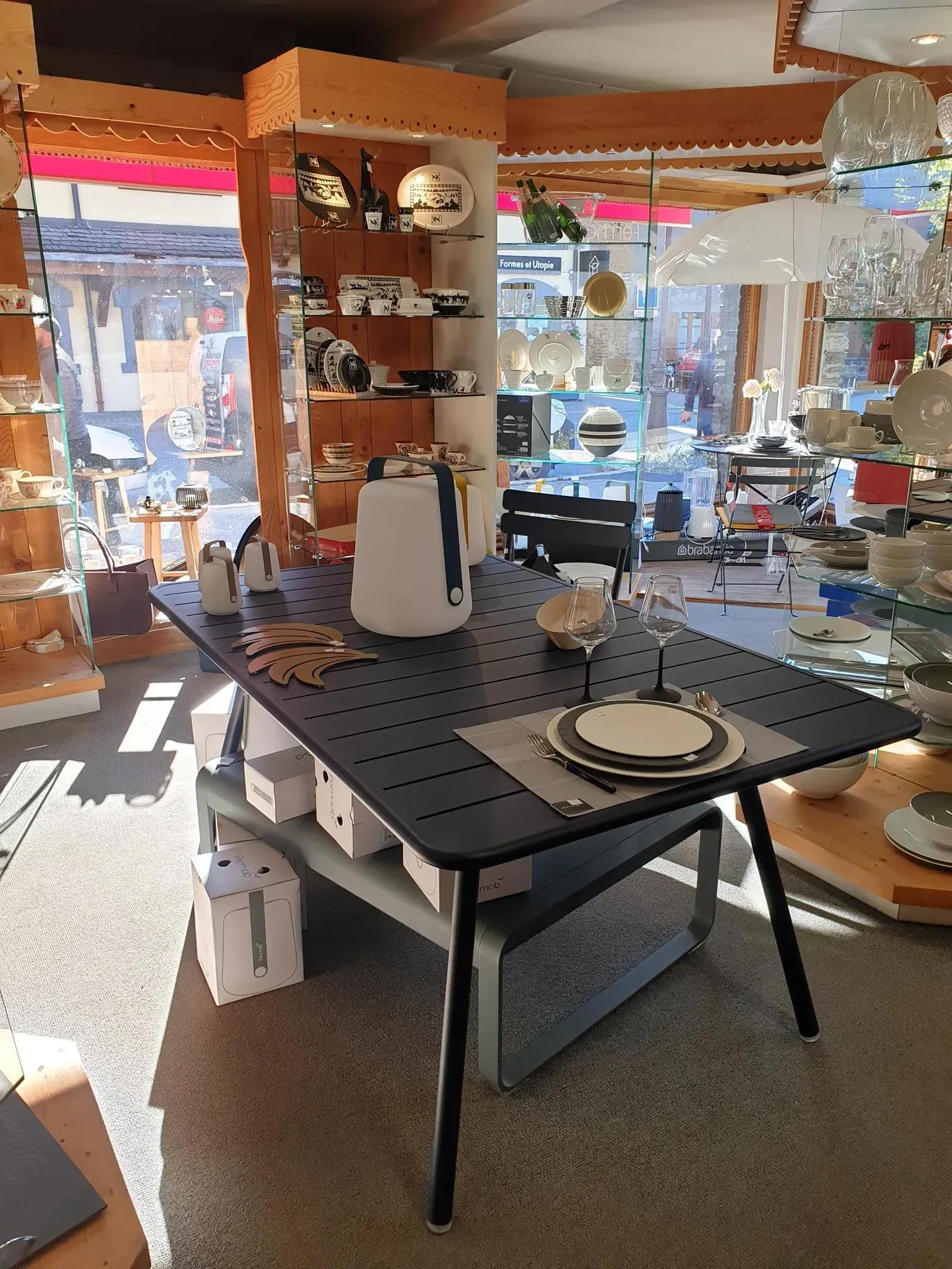 Table d'exposition dans un magasin de décoration intérieure avec vaisselle, vases et suspensions lumineuses près d'une grande fenêtre.