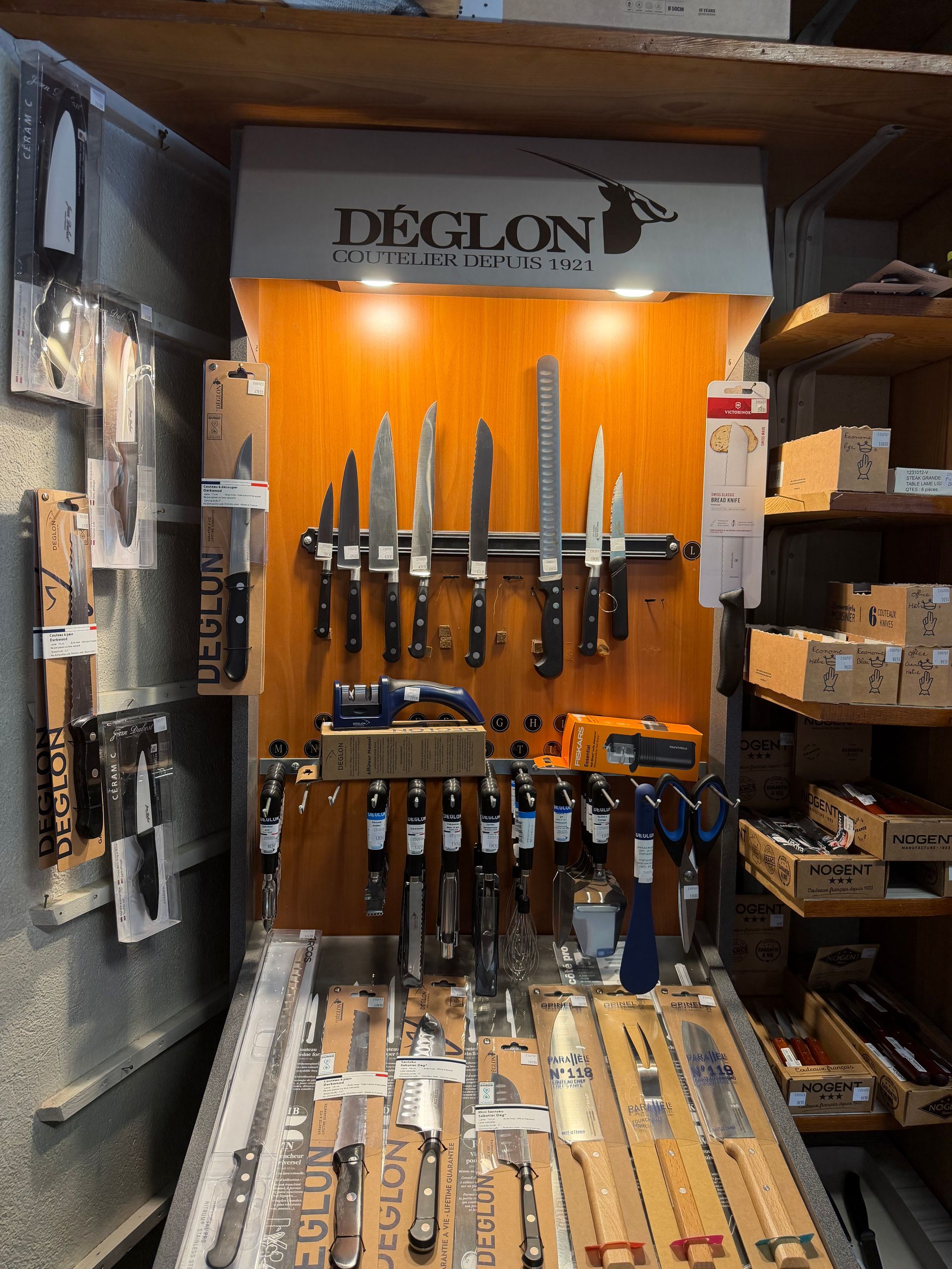 Présentoir à couteaux et outils dans un magasin, avec des lames fixées sur un mur en bois et des étagères de boîtes sur les côtés.