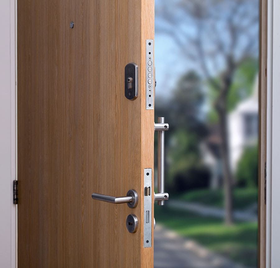 Porte d’entrée en bois entrouverte, laissant voir la poignée, la serrure et le paysage extérieur flou au-delà.