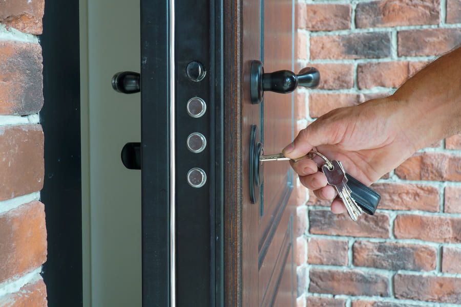 Main déverrouillant une porte d’entrée noire à l’aide de clés, à côté d’un mur en briques