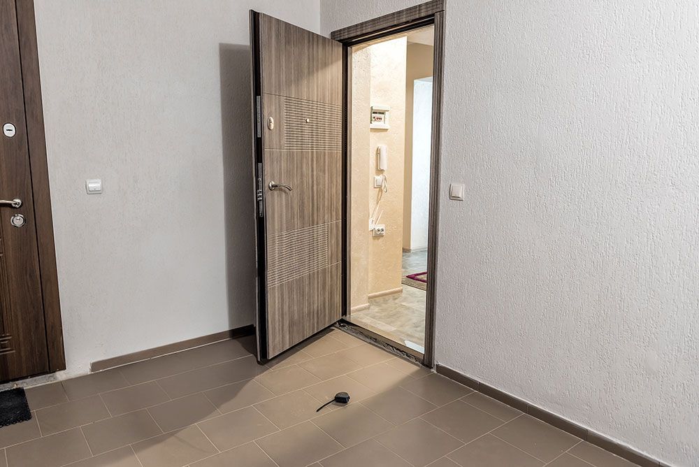 Porte d’appartement ouverte sur un couloir éclairé, avec une pièce beige et un sol marron au premier plan.
