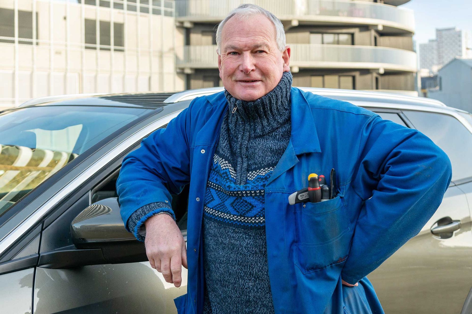 Un homme appuyé contre une voiture devant un bâtiment, portant une veste bleue et souriant.