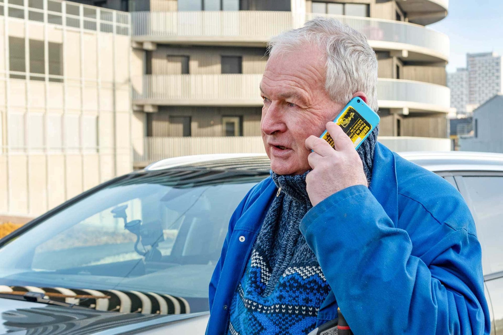 Un homme tenant un téléphone jaune à côté d’une voiture, devant un bâtiment moderne