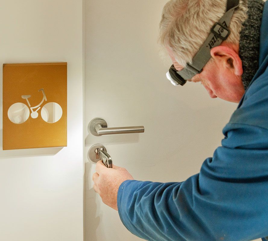 Un homme portant une lampe frontale répare une serrure de porte près d’une plaque murale dorée dans un couloir.