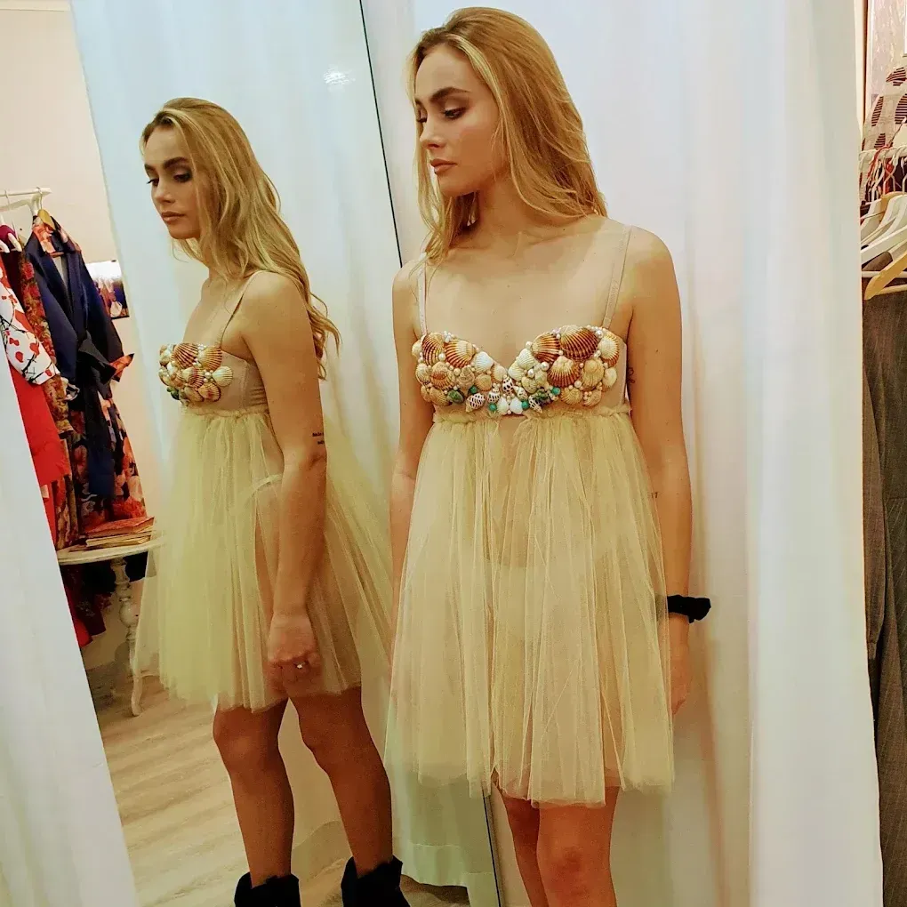 Una mujer con un vestido adornado con conchas marinas está frente a un espejo en una tienda de ropa.
