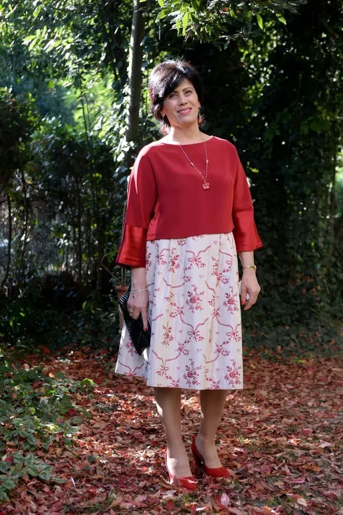 Una mujer con una blusa roja y una falda blanca está parada entre las hojas.