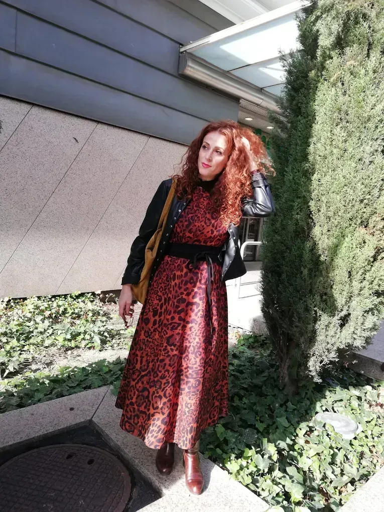 Mujer con un vestido rojo estampado y una chaqueta negra parada afuera, tocándose su cabello rojo y rizado.