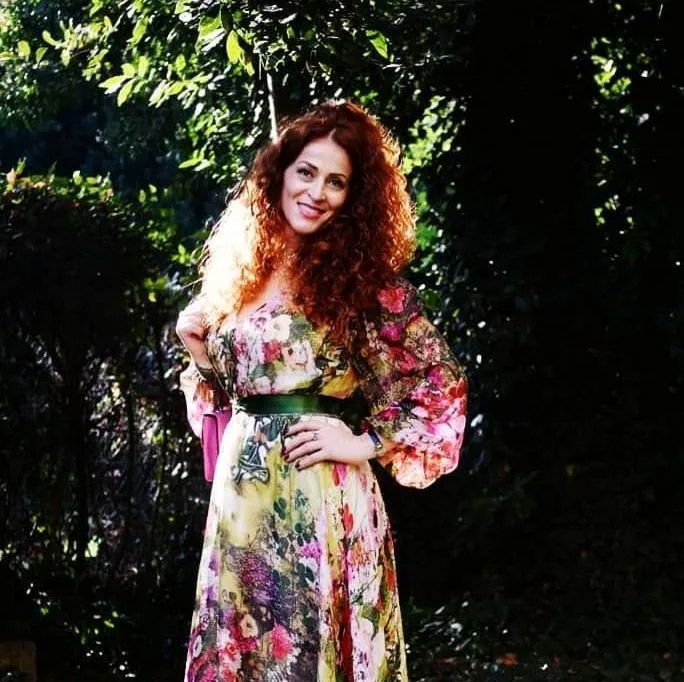 Mujer con vestido floral y cabello rojo y rizado sonríe al aire libre, con la mano en la cadera.