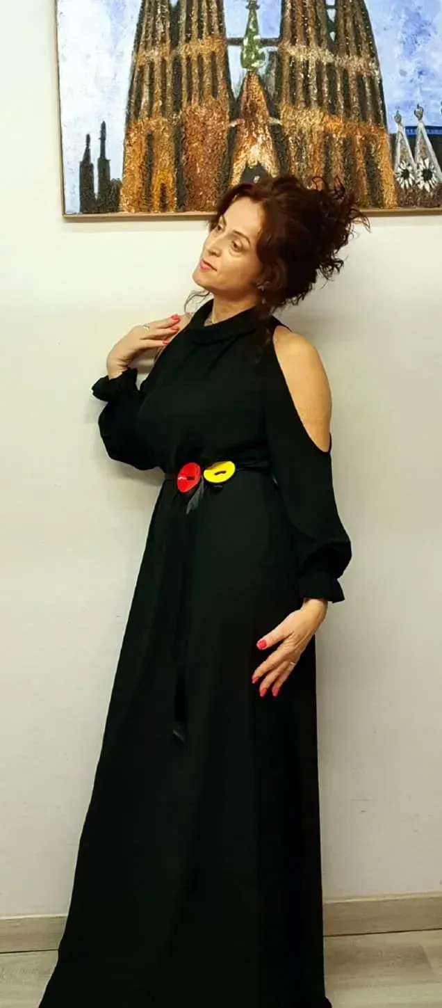 Una mujer con un vestido negro, mangas de hombros descubiertos y un cinturón colorido se encuentra frente a una pared con obras de arte.