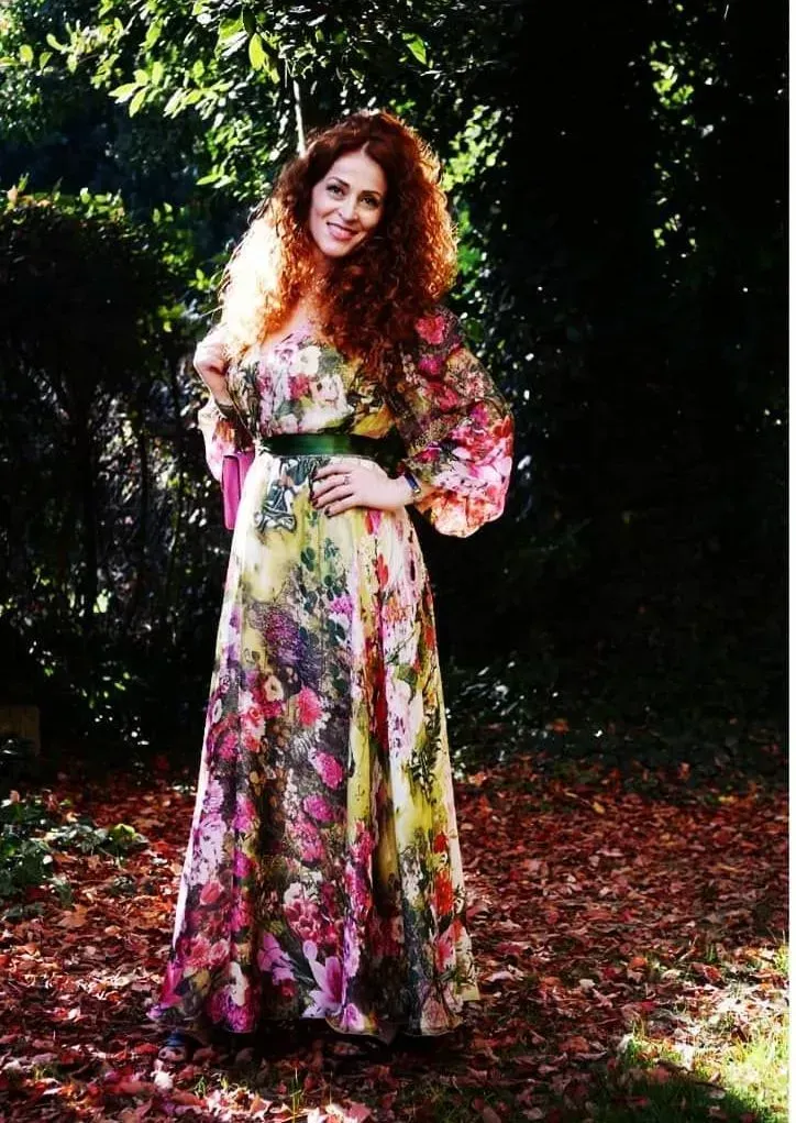 Mujer con vestido floral posa al aire libre, cabello rojo y rizado, cinturón verde, follaje soleado.