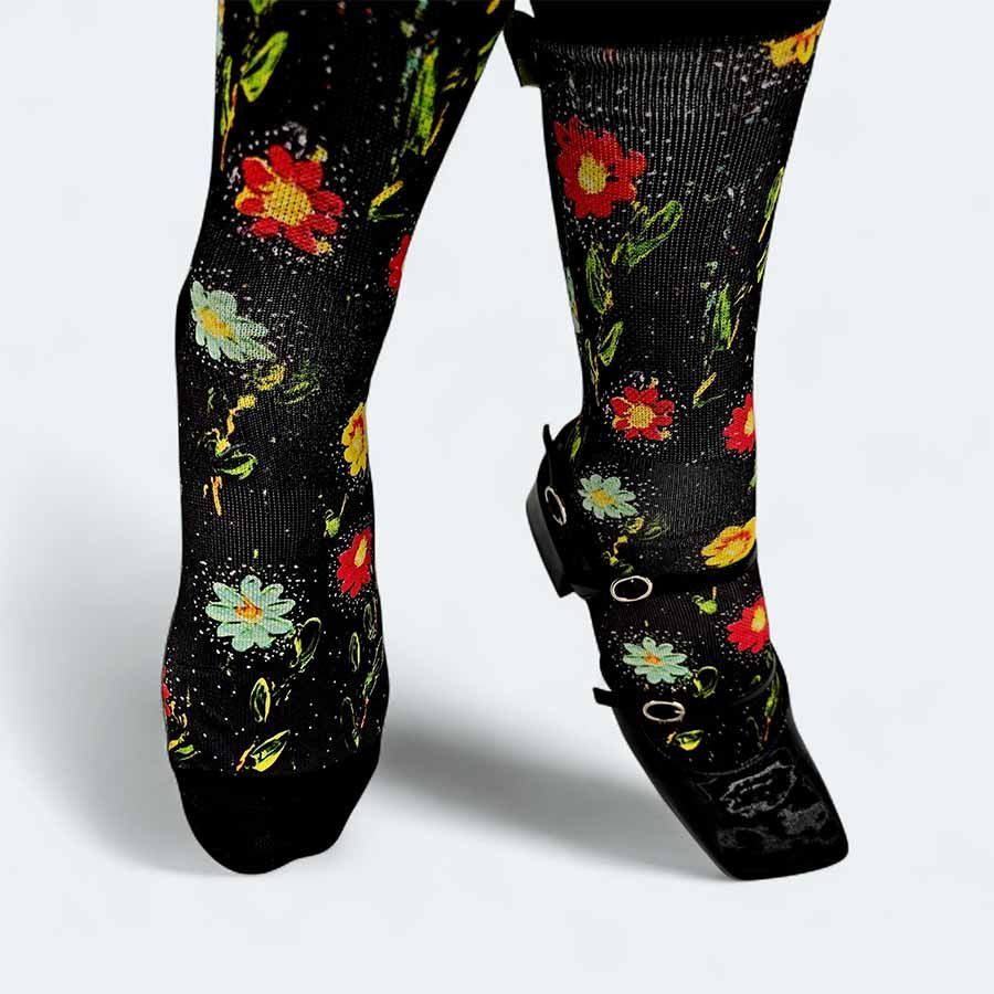 Pies con calcetines negros con estampado floral y zapatos negros de punta cuadrada, posando sobre un fondo blanco.