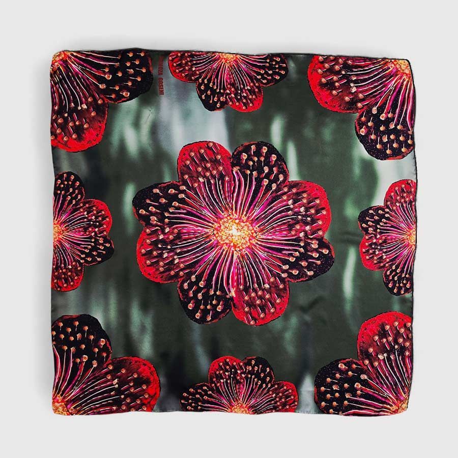 Bufanda cuadrada con diseño floral abstracto repetido en rojo y negro sobre un fondo verde apagado.