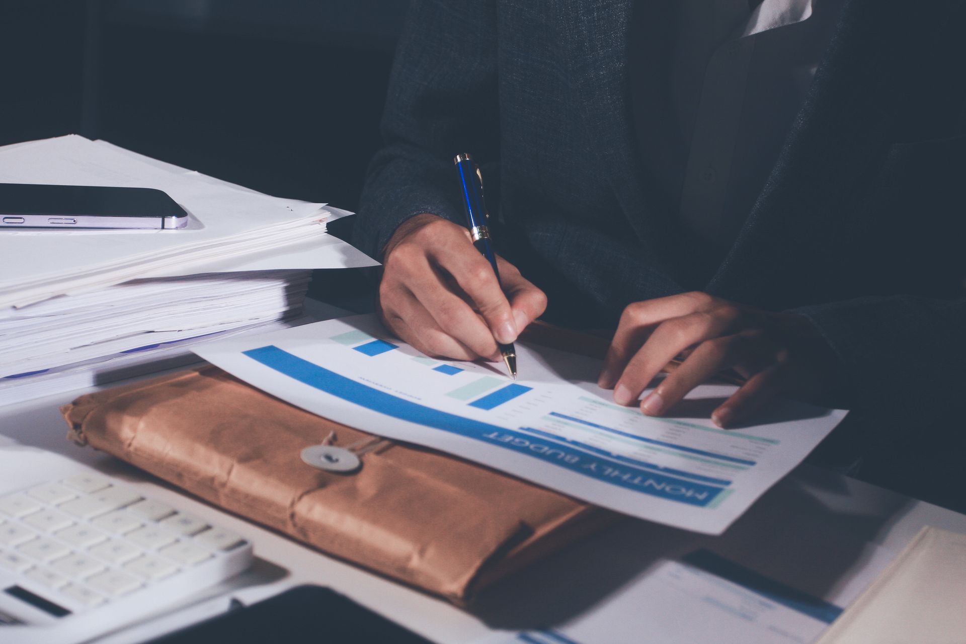 Une personne en costume signe des documents à un bureau avec des graphiques et un porte-documents en cuir marron.
