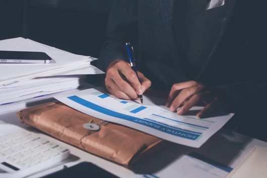 Une personne en costume signe des documents à un bureau avec des graphiques et un porte-documents en cuir marron.