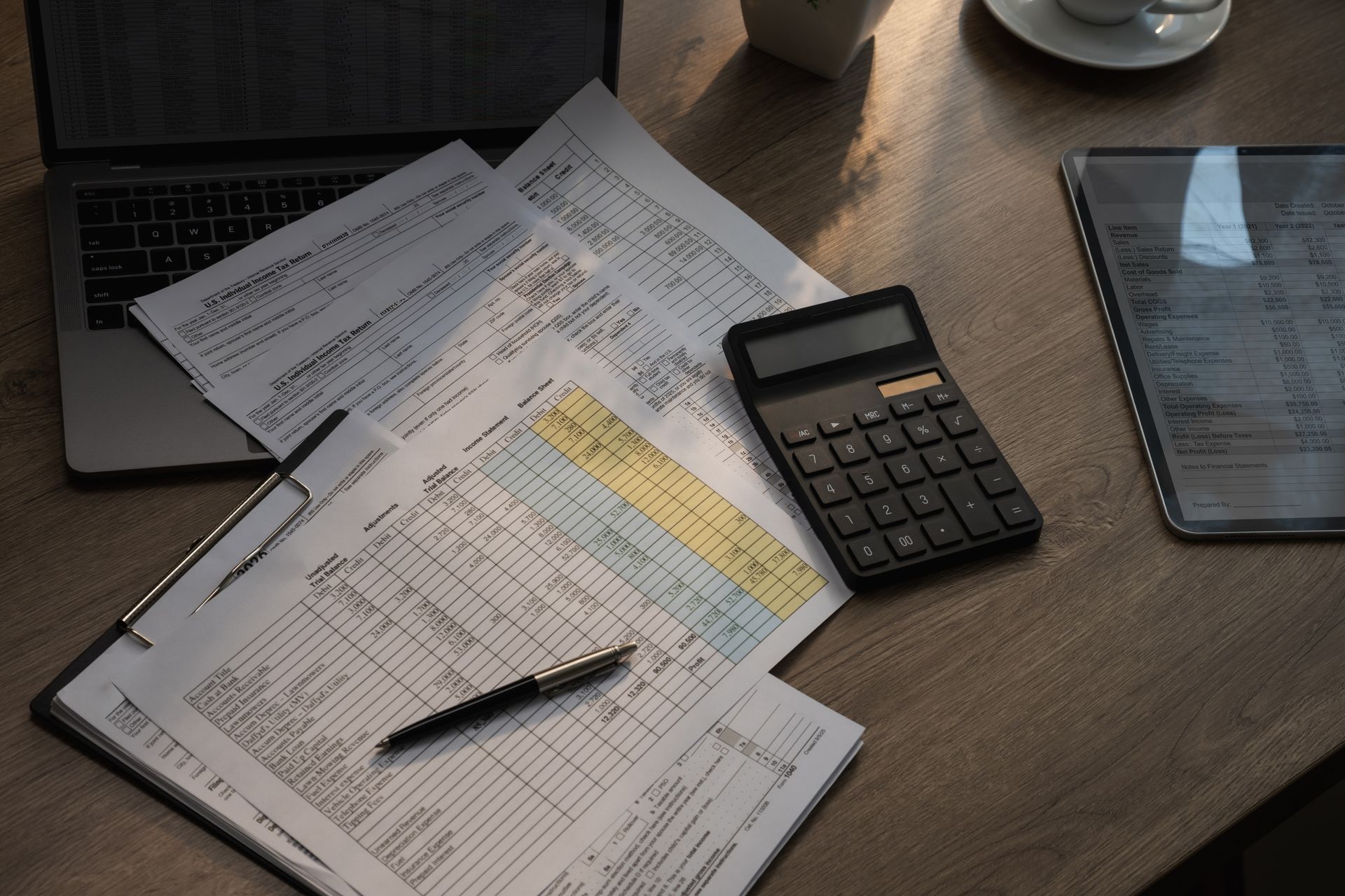 Une calculatrice et des papiers éparpillés sur un bureau, avec un stylo et un ordinateur portable en arrière-plan.