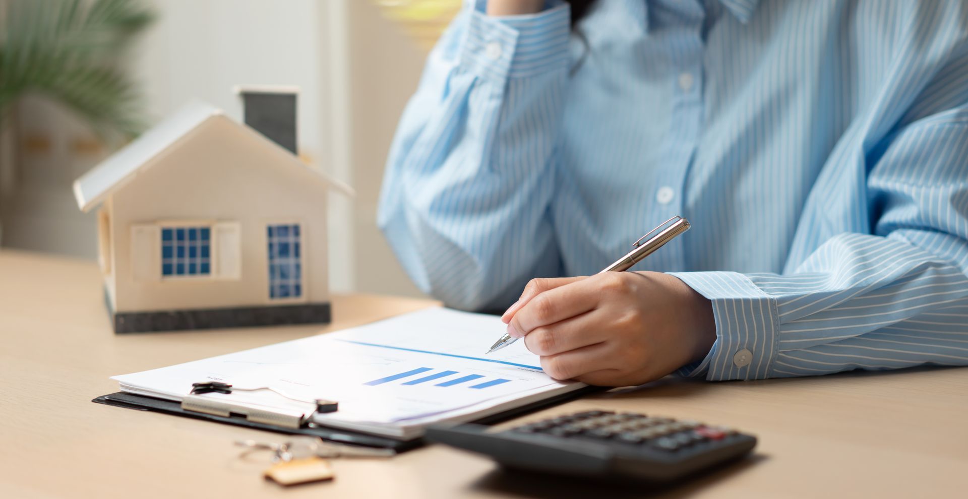 Des mains écrivent sur des documents financiers à côté d'une calculatrice et d'une maquette de maison posés sur un bureau.