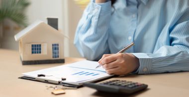 Des mains écrivent sur des documents financiers à côté d'une calculatrice et d'une maquette de maison posés sur un bureau.