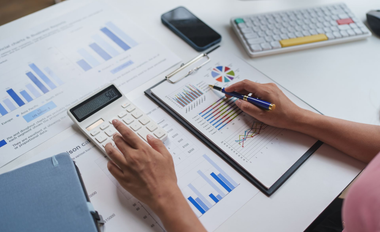 Des mains tenant une calculatrice et un stylo consultent des graphiques sur un bloc-notes à un bureau.