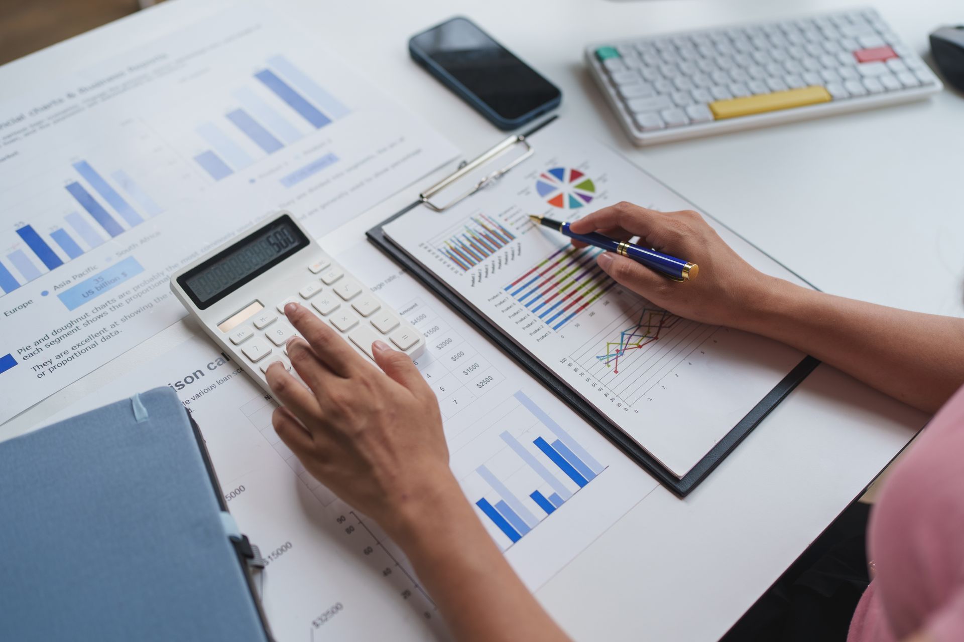 Des mains utilisant une calculatrice et un stylo au-dessus de graphiques financiers posés sur un bureau où se trouvent un ordinateur portable, un téléphone et un clavier.