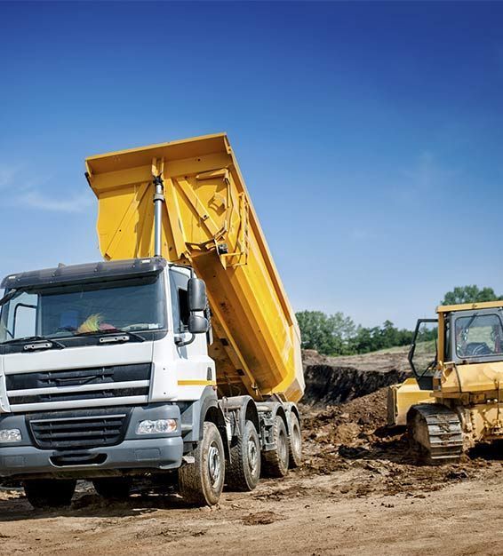 Un camión volquete amarillo con la caja elevada junto a una excavadora amarilla en una obra de construcción de tierra bajo un cielo azul.
