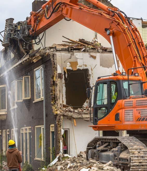 Una excavadora naranja derriba un edificio de ladrillos mientras un trabajador rocía agua en el lugar de la demolición para controlar el polvo.