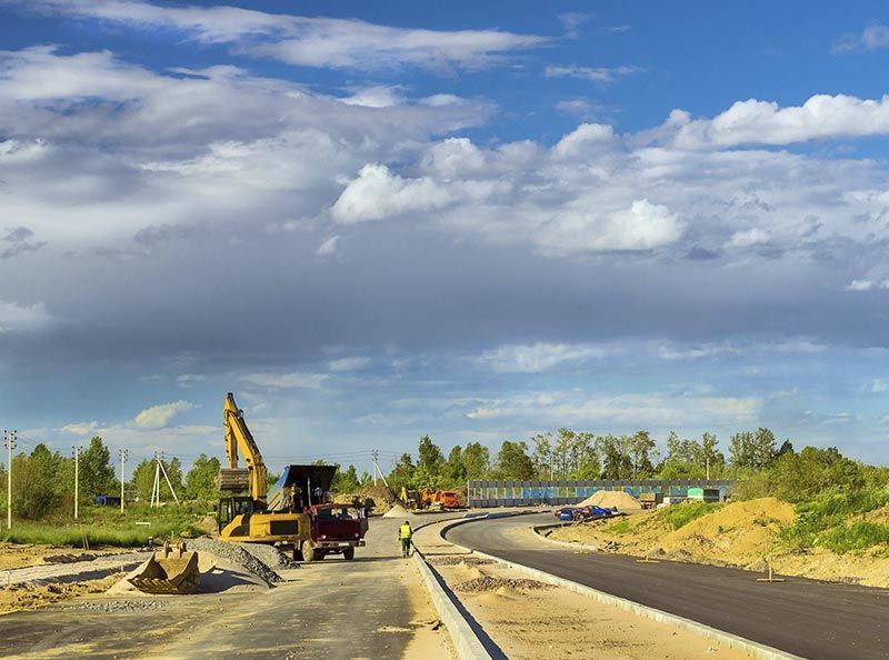 En una obra de construcción se observa una excavadora trabajando en una carretera pavimentada bajo un cielo parcialmente nublado.