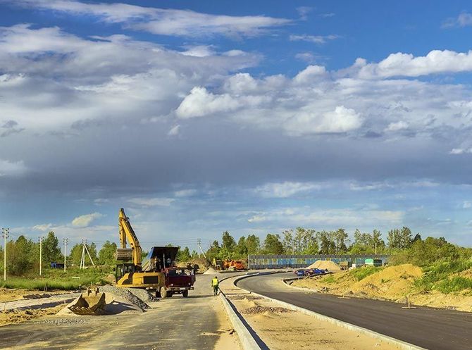 En una obra de construcción se observa una excavadora trabajando en una carretera pavimentada bajo un cielo parcialmente nublado.