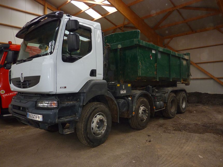 Un camion-benne blanc avec un conteneur de fret vert est stationné à l'intérieur d'un grand entrepôt aux poutres en bois.