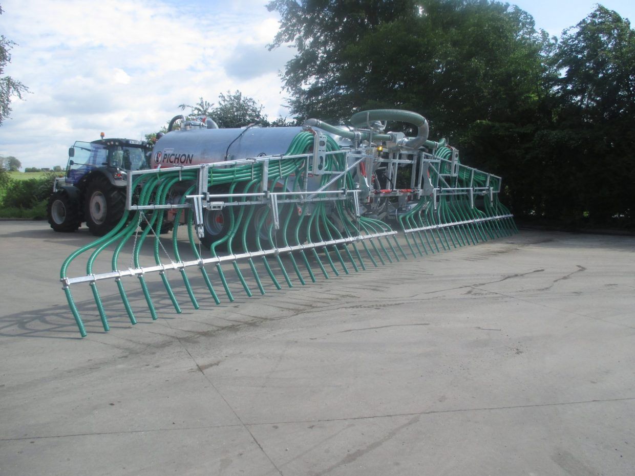 Un tracteur tirant une grande citerne équipée d'une large rampe d'injection de lisier agricole à plusieurs tubes sur un terrain extérieur en béton.
