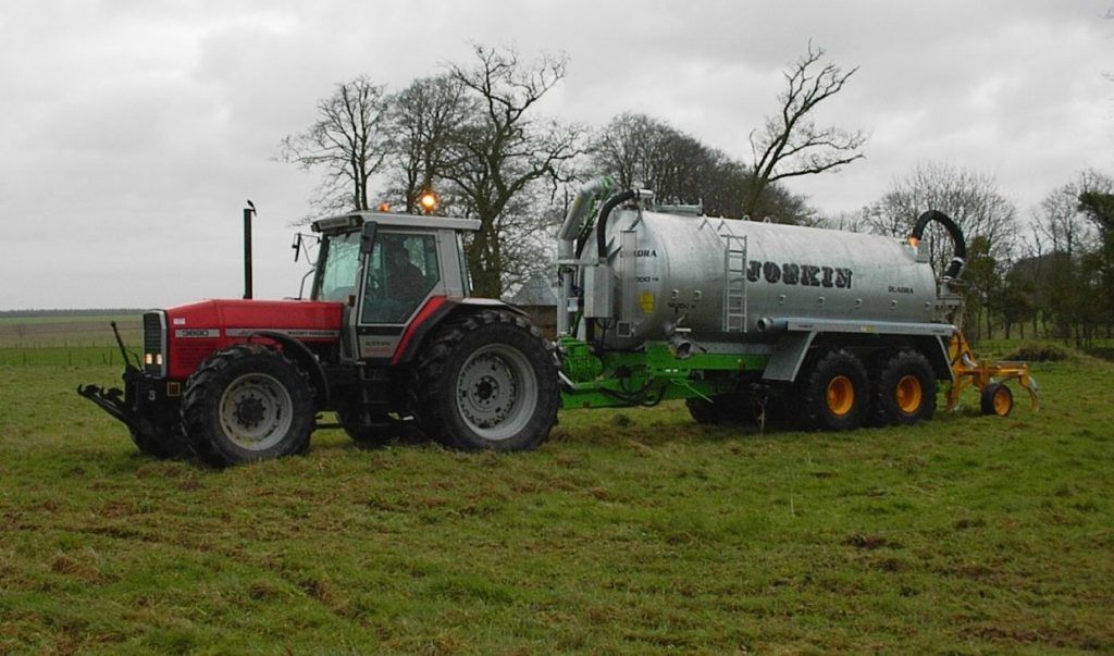 Un tracteur rouge tirant une citerne agricole argentée à travers un champ herbeux par temps couvert.