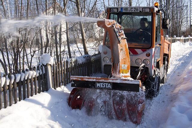 Une souffleuse à neige orange dégage la neige d'un chemin longeant une clôture en bois par une journée d'hiver ensoleillée.