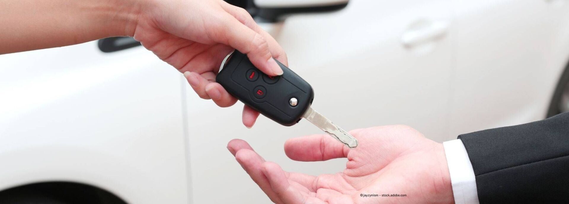 Vor einem weißen Auto wird ein Autoschlüssel von einer Person an eine andere übergeben.