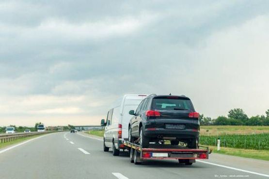 Ein weißer Lieferwagen zieht einen Anhänger mit einem schwarzen SUV auf einer Autobahn unter einem bewölkten Himmel.