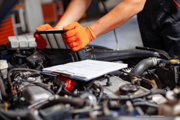 Mechaniker mit orangefarbenen Handschuhen tauscht einen Luftfilter in einem Automotor aus, in der Nähe ein Klemmbrett.