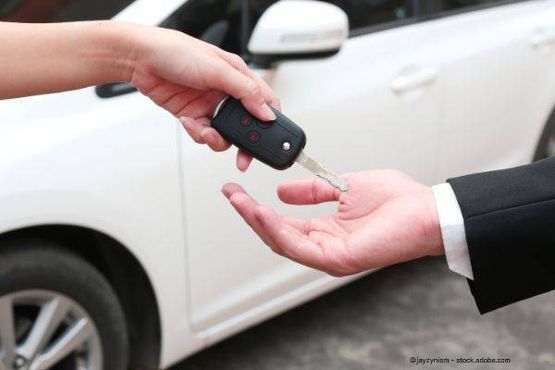 Person übergibt Autoschlüssel an andere Person; weißes Auto im Hintergrund.
