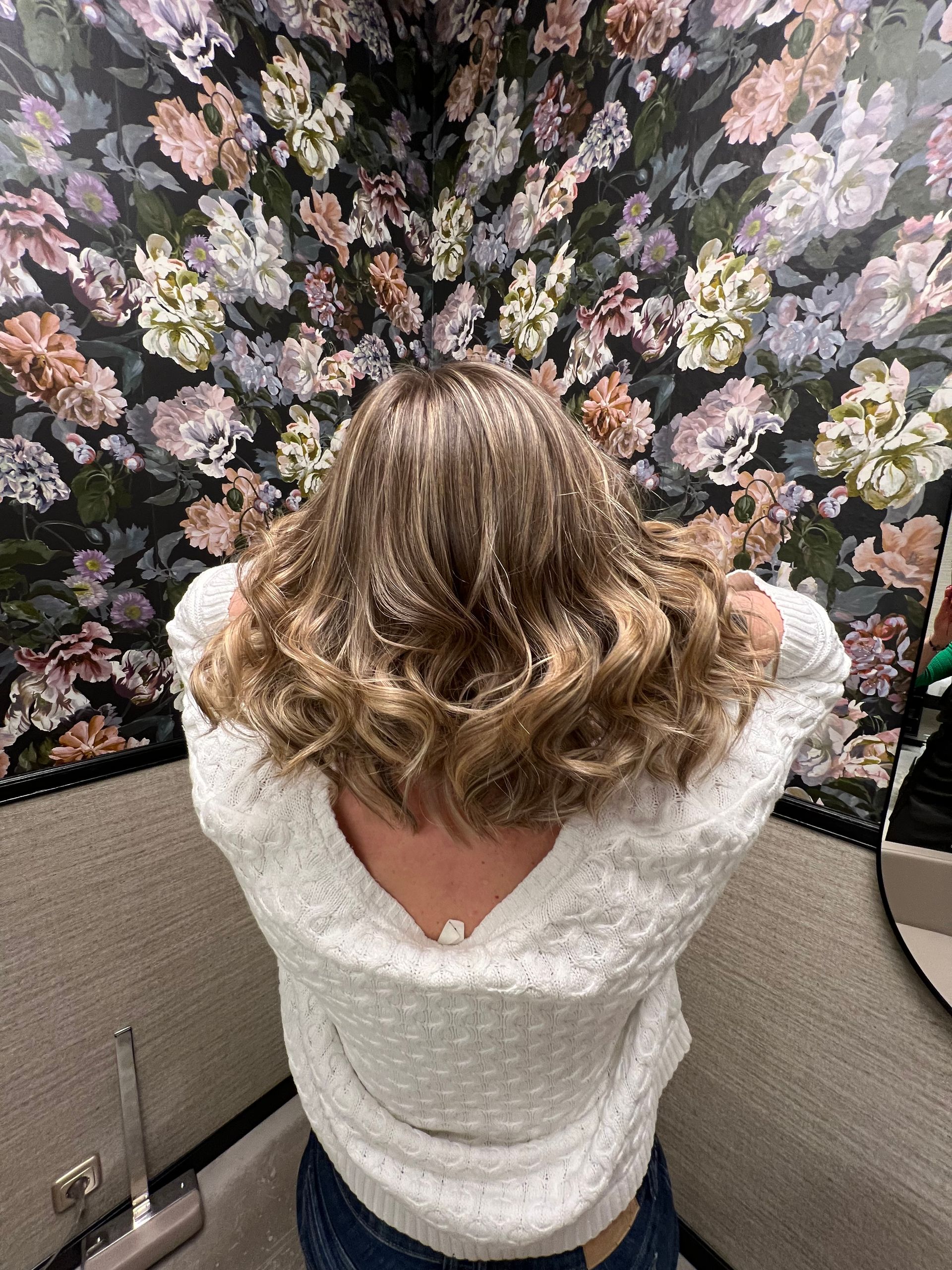 Frau mit gewelltem braunem Haar, die einen weißen Pullover trägt, in einem Zimmer mit geblümter Tapete.