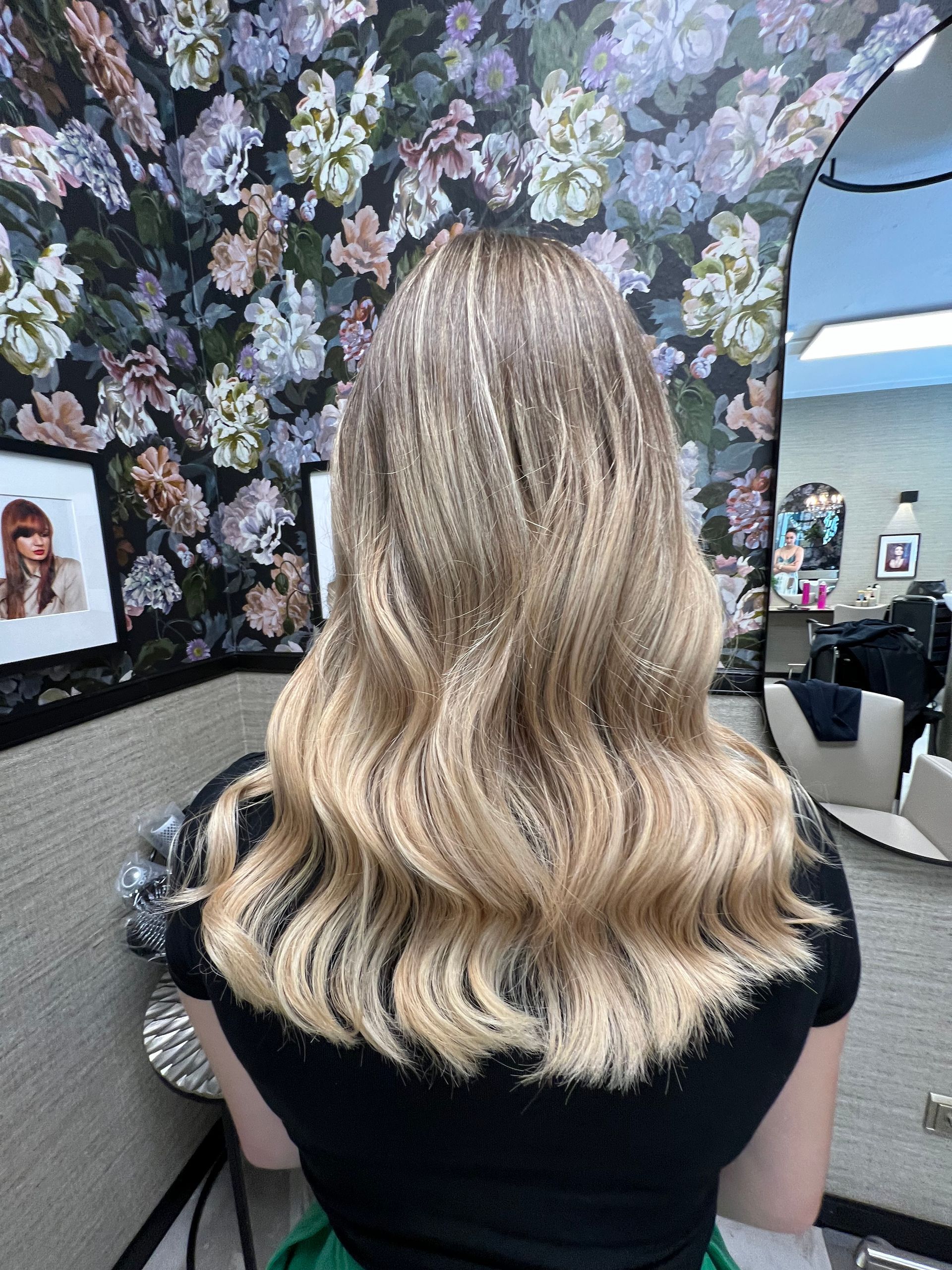Frau mit langem, gewelltem blonden Haar in einem Salon vor einer floralen Tapetenkulisse.