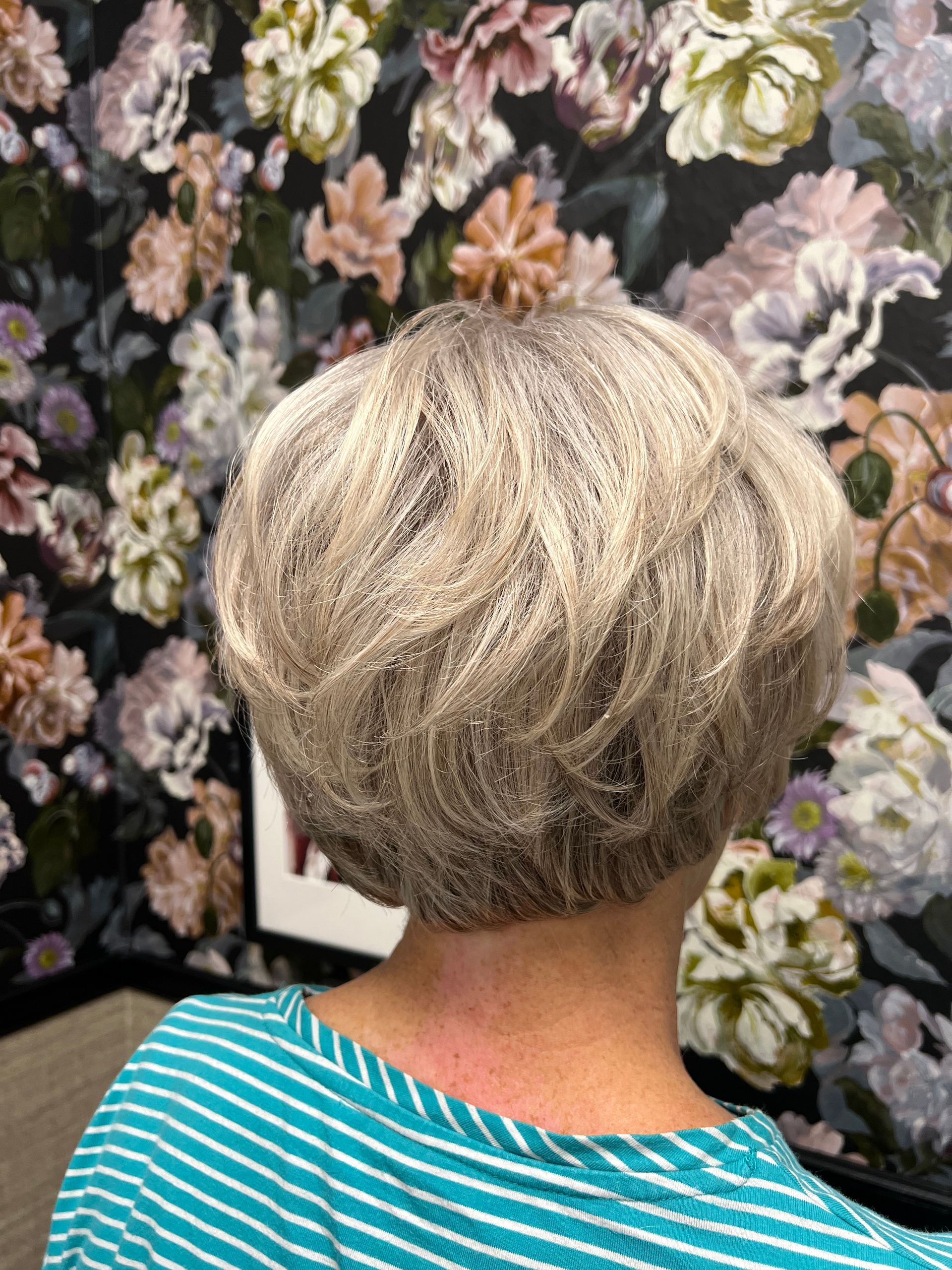 Kurzes, gestuftes blondes Haar in einer abgerundeten Form. Floraler Tapetenhintergrund.