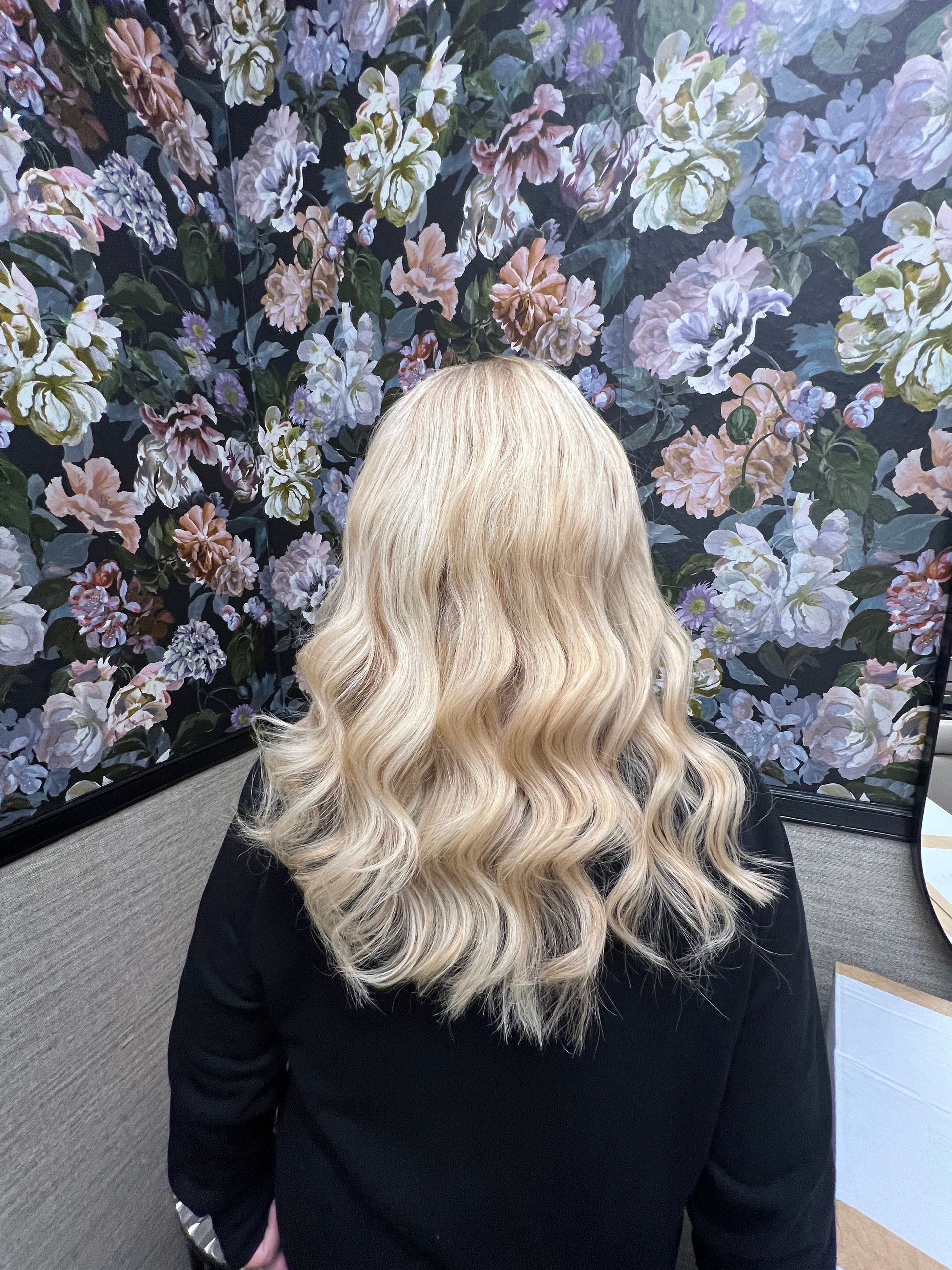 Blondes, gewelltes Haar vor einem floralen Tapetenhintergrund.