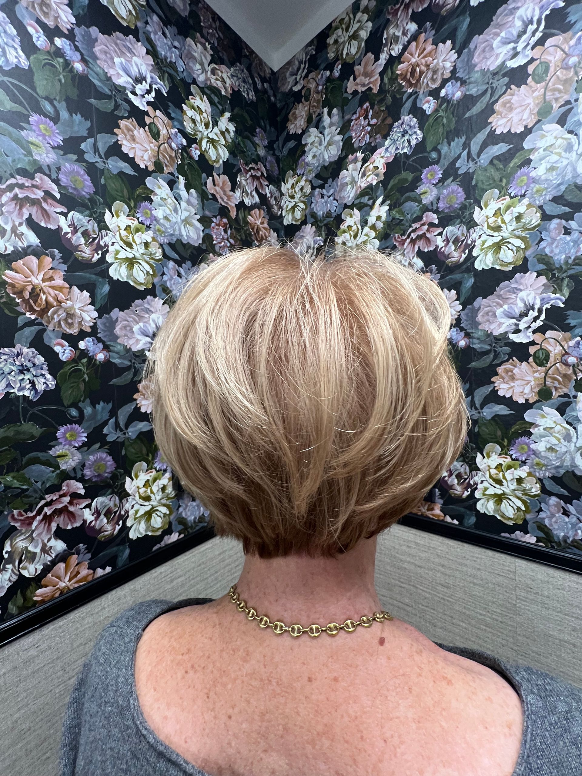 Frau mit kurzen blonden Haaren und einer goldenen Halskette vor einer geblümten Tapete.