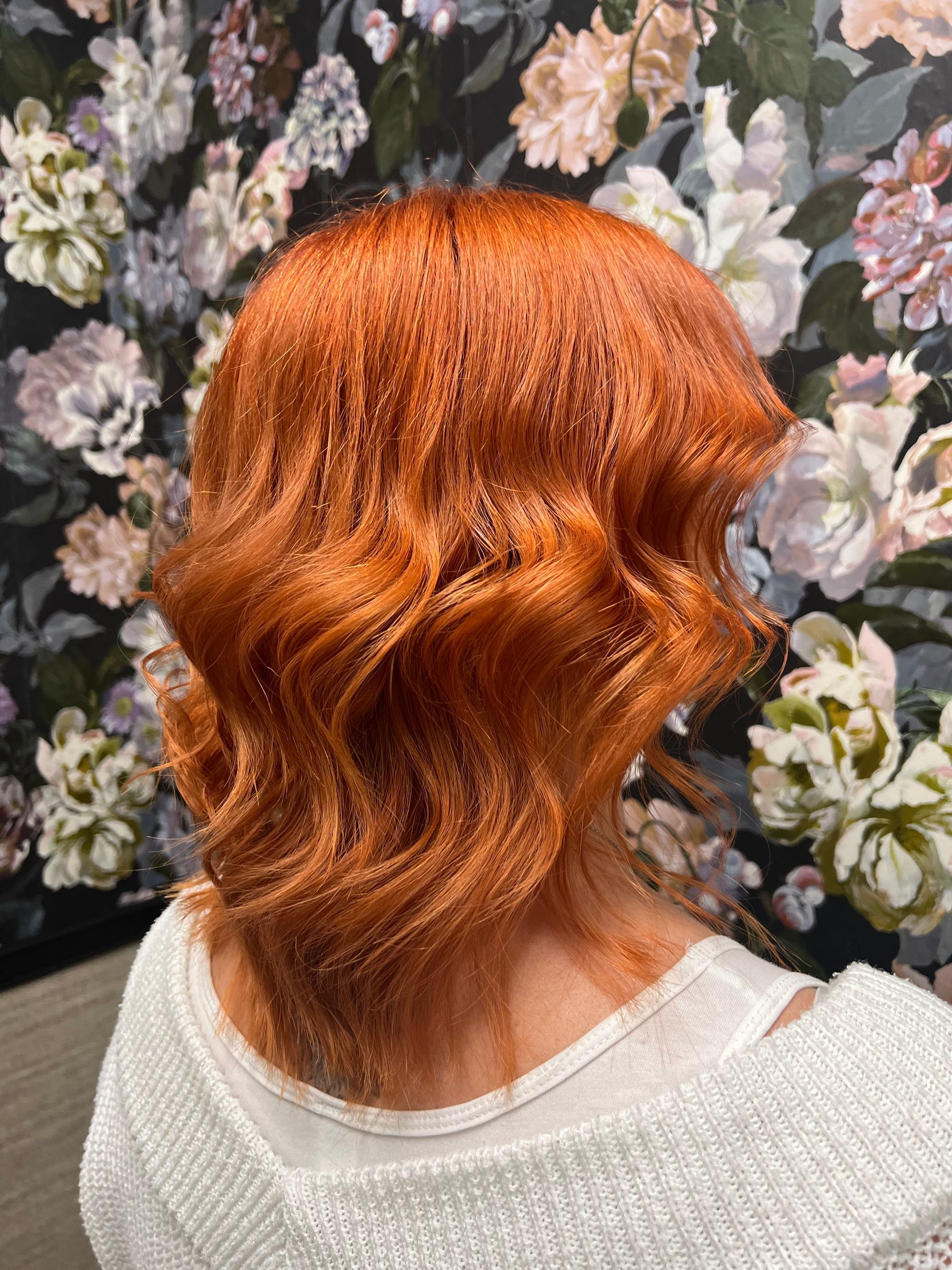 Frau mit leuchtend rotem, welligem Haar vor geblümter Tapete; trägt einen weißen Pullover.