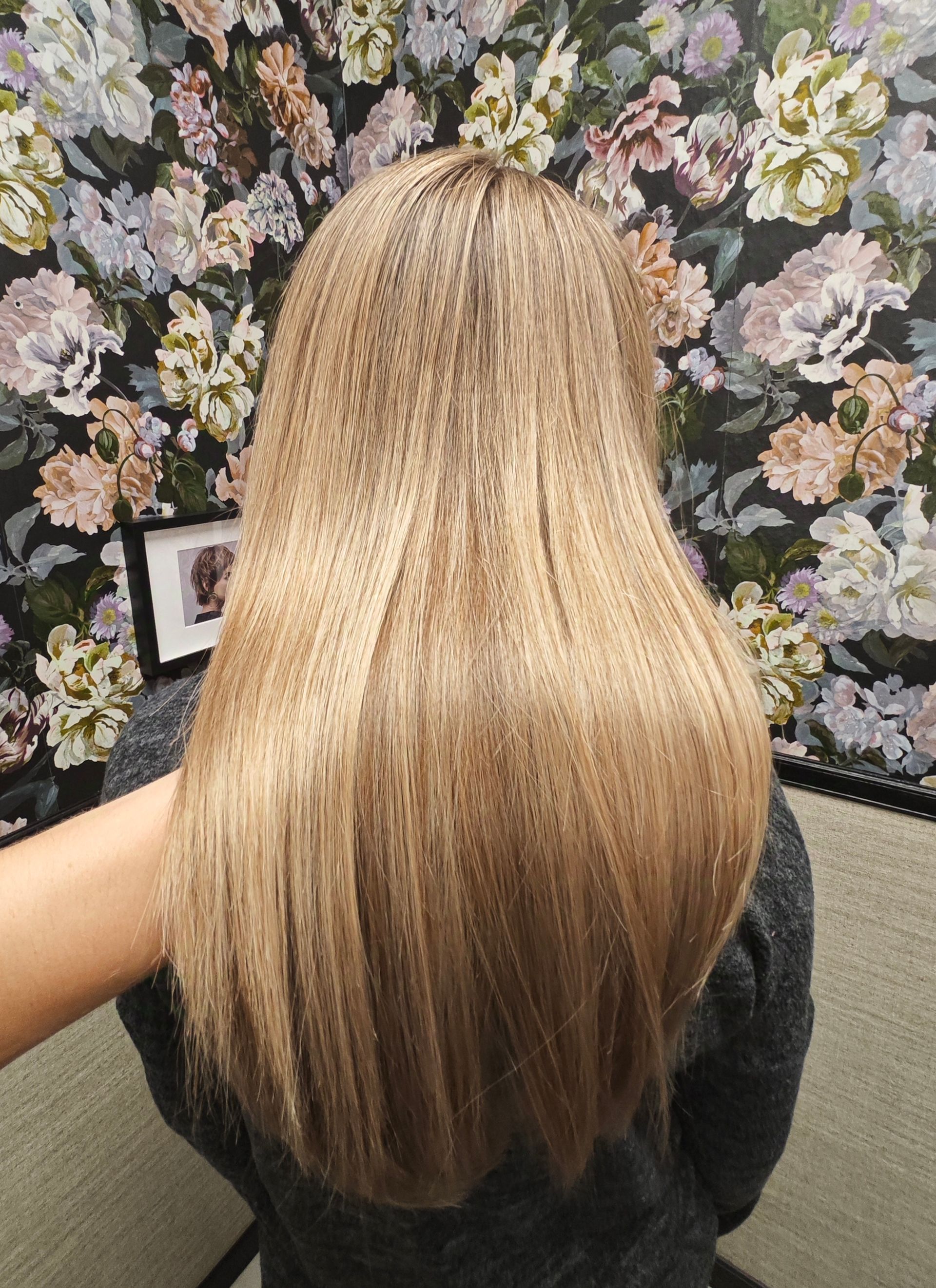 Blondes, gekräuseltes Haar, das den Rücken einer Person hinunterläuft, dunkles Oberteil, floraler Hintergrund.