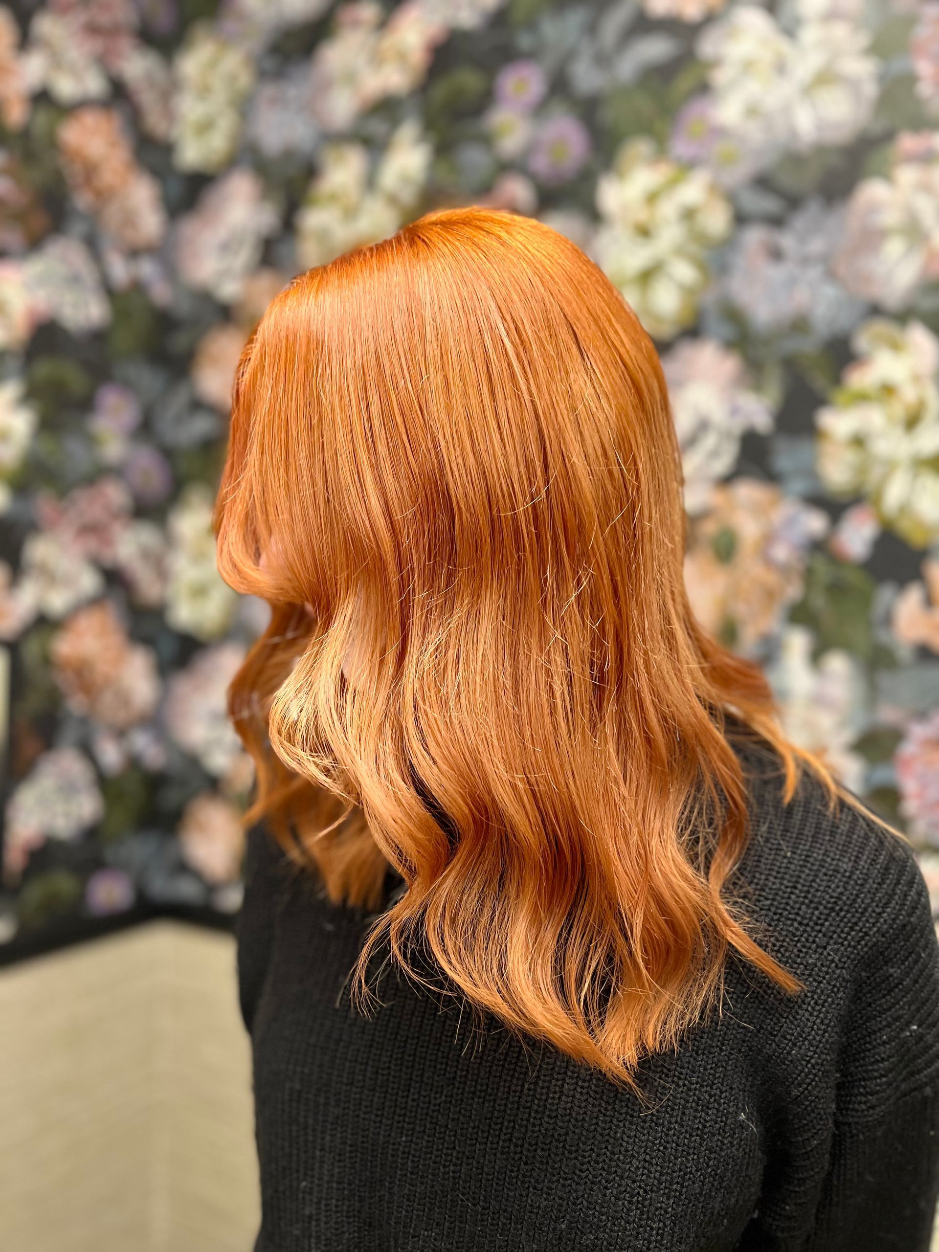 Frau mit leuchtend kupferfarbenem Haar. Wellige Frisur, schwarzer Pullover, florale Tapete im Hintergrund.
