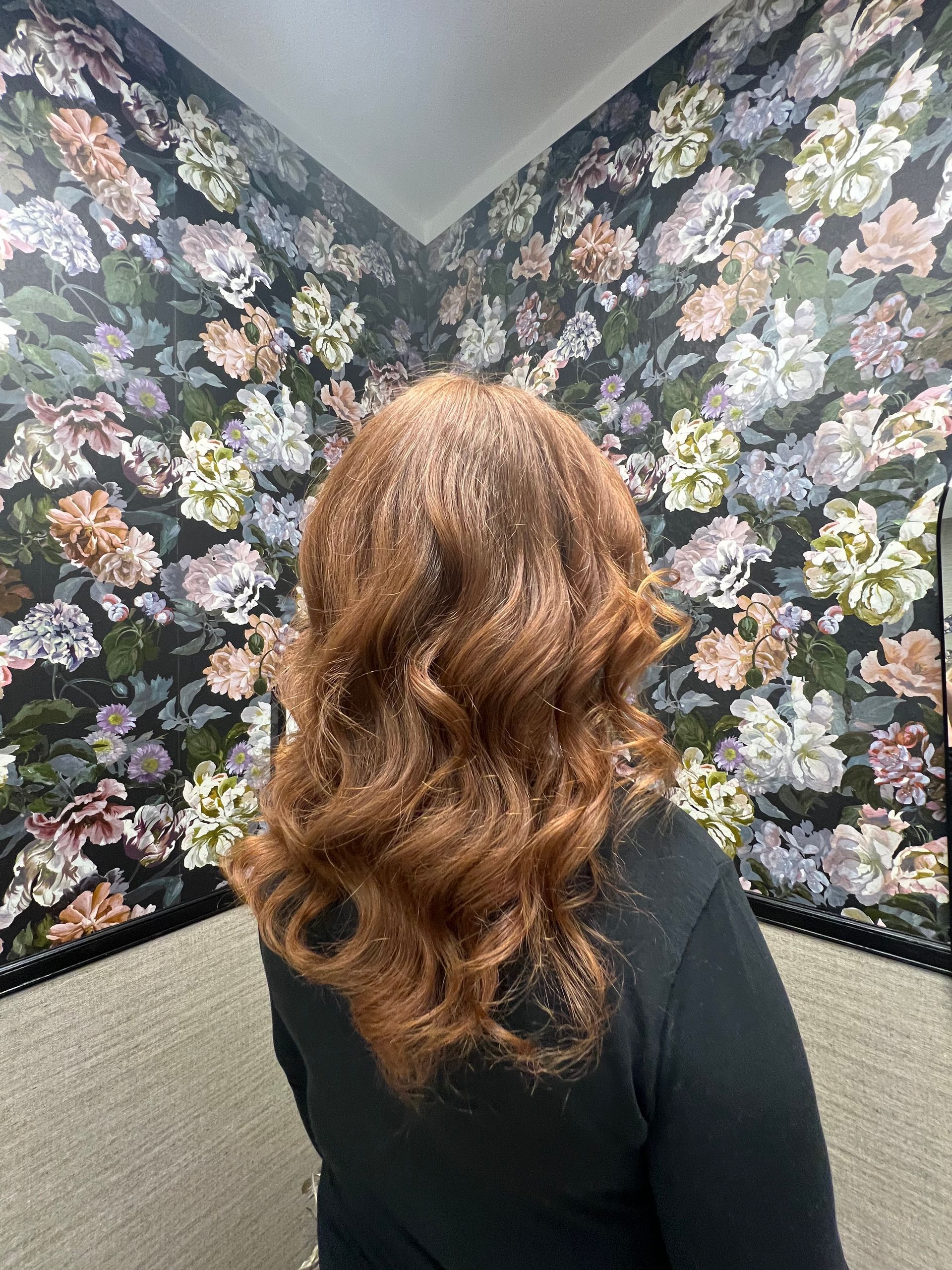 Eine Frau mit kupferfarbenem, gewelltem Haar steht in einer Ecke vor einer floralen Tapete und einem neutralfarbenen Teppich.