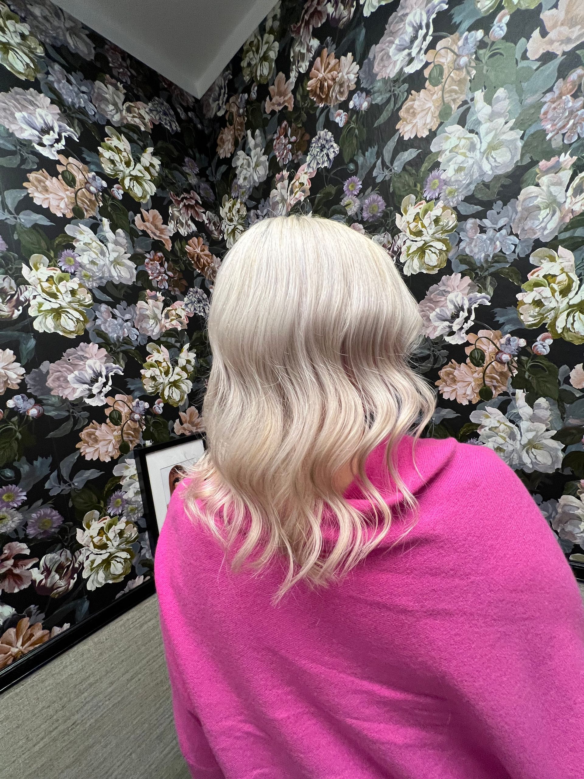 Blondes, gewelltes Haar, rosa Pullover, geblümter Tapetenhintergrund.