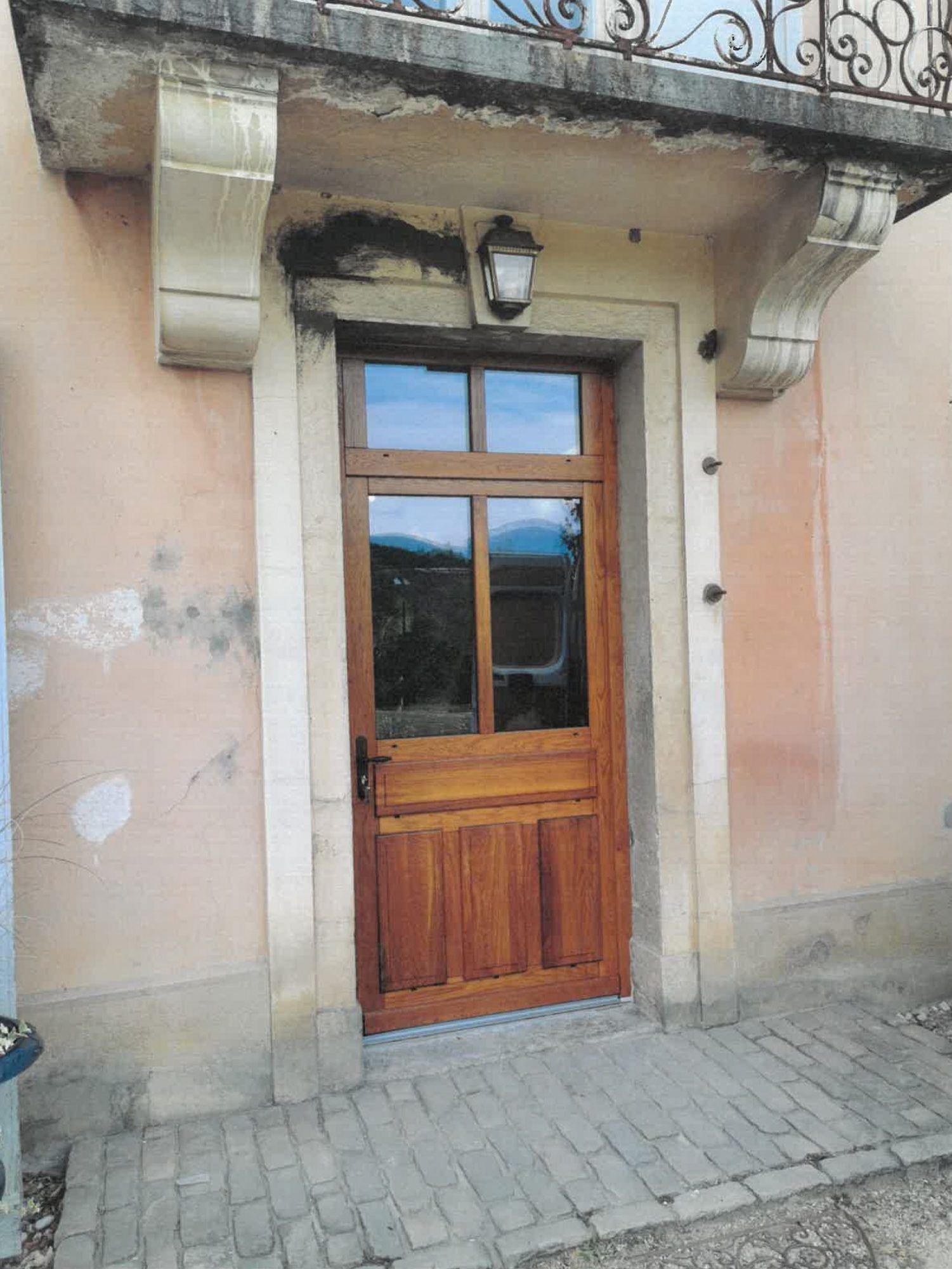 Porte en bois avec panneau de verre, sous un linteau en pierre et un balcon, sur un bâtiment rose avec allée en briques.