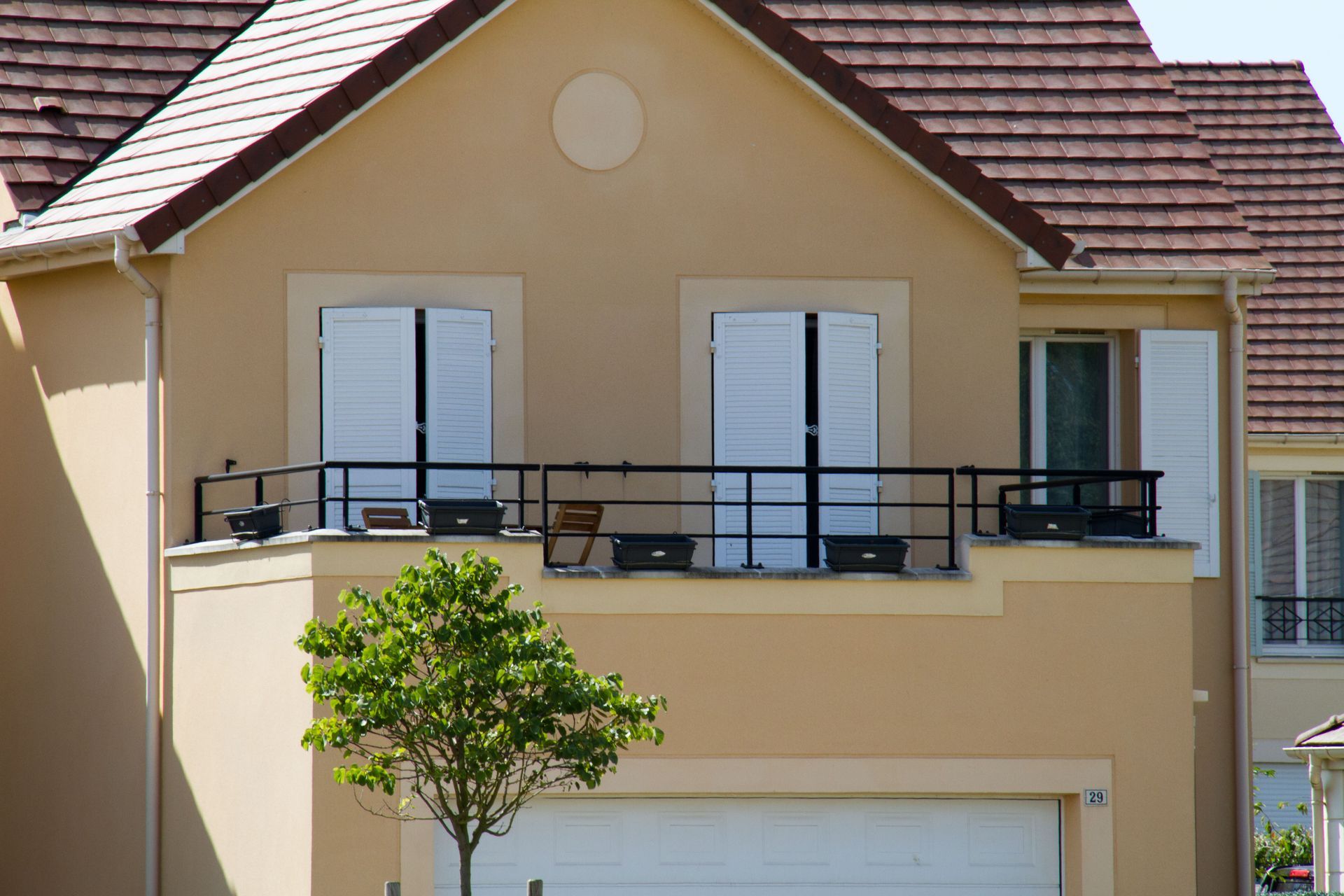 Maison beige avec balcon, volets blancs et toit marron. Un petit arbre se trouve devant.