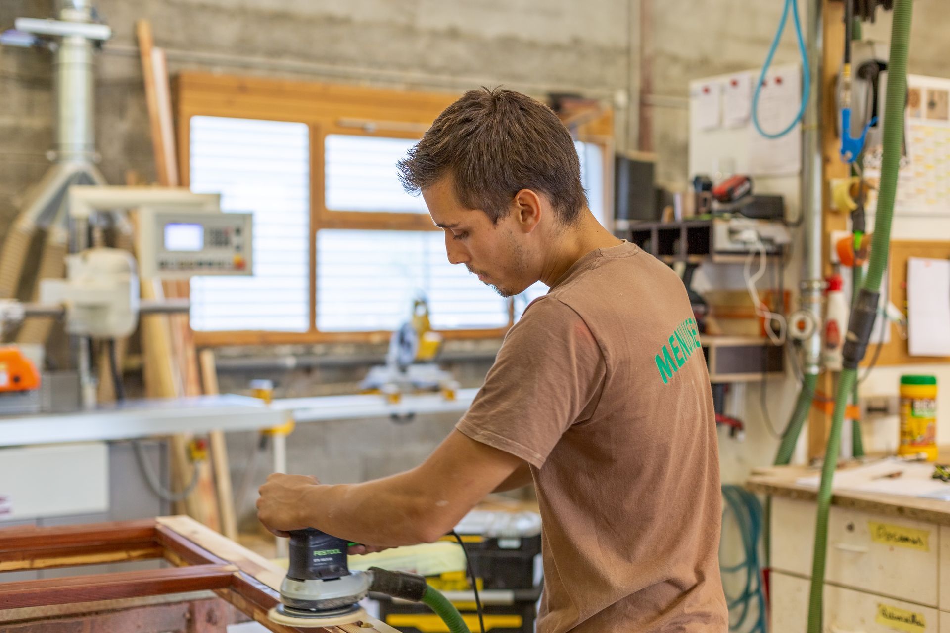 Un menuisier ponçant du bois dans un atelier, vêtu d'une chemise marron.